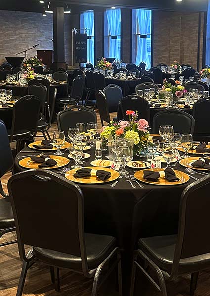 Elegant banquet tables set for steak and cocktails, with gold plates, black napkins, and florals.