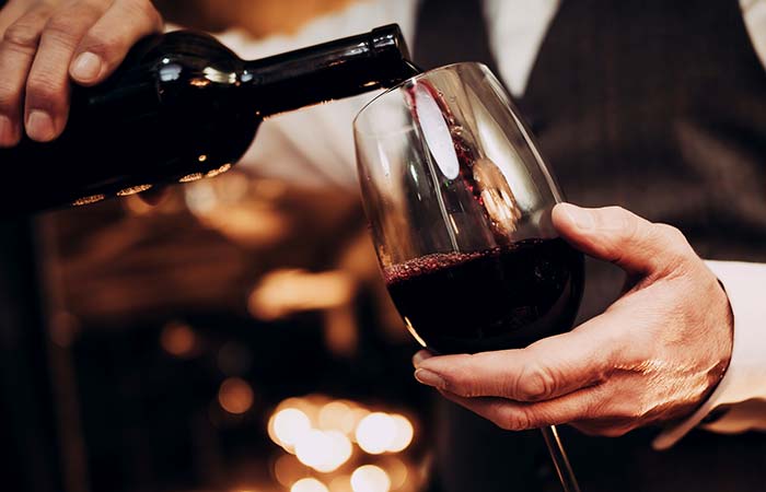 Person pouring red wine into a glass, dressed in a vest and white shirt, preparing for steak.