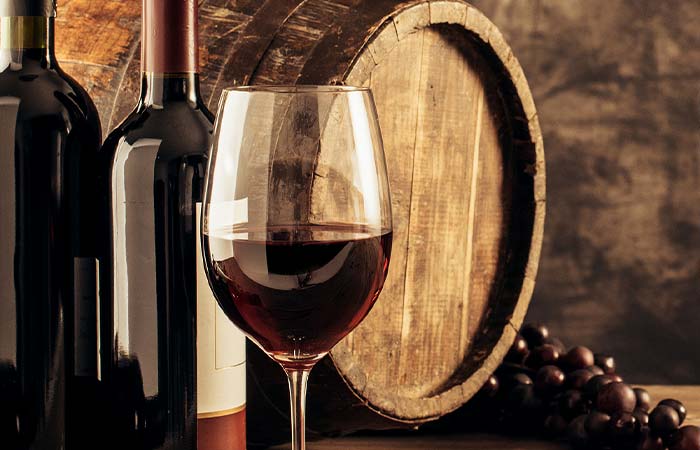 A glass of red wine, steak, two wine bottles, grapes, and a wooden barrel in the background.